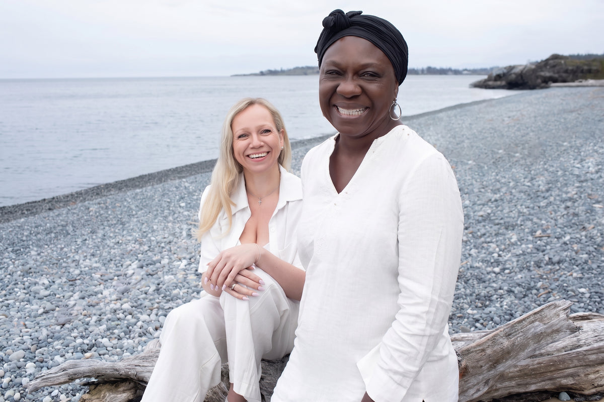 Two women in white linen laughing together on a pebble beach by the sea