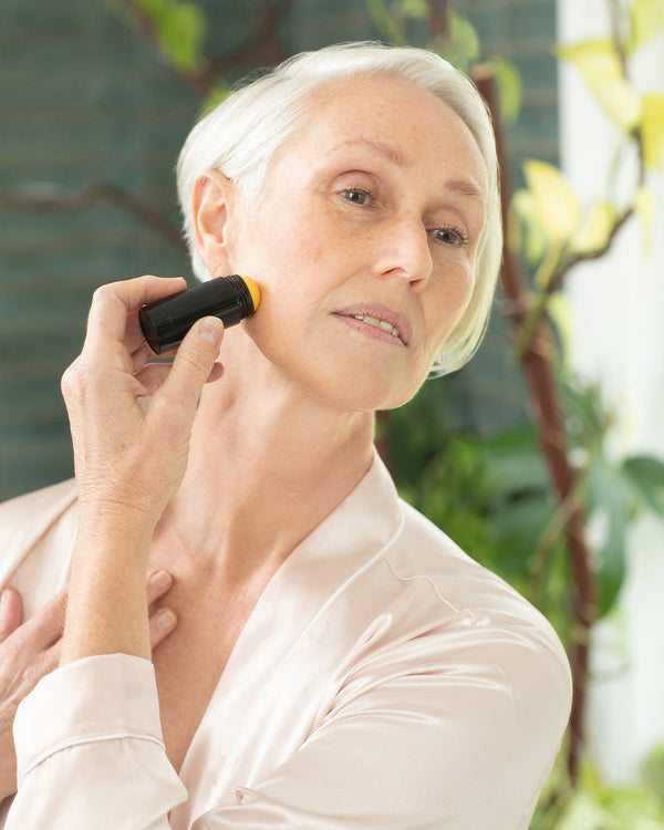 Barbi gliding the PHILOGENI Resilience Balm stick across her cheekbone in soft morning light.